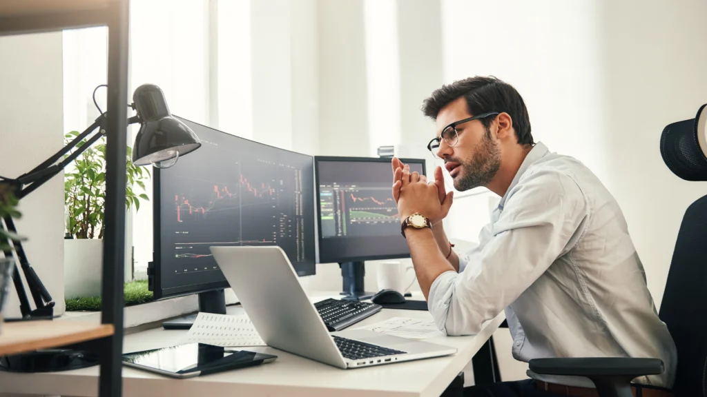 man looking at laptop and computer monitors with cfd trading charts on them, while sitting down