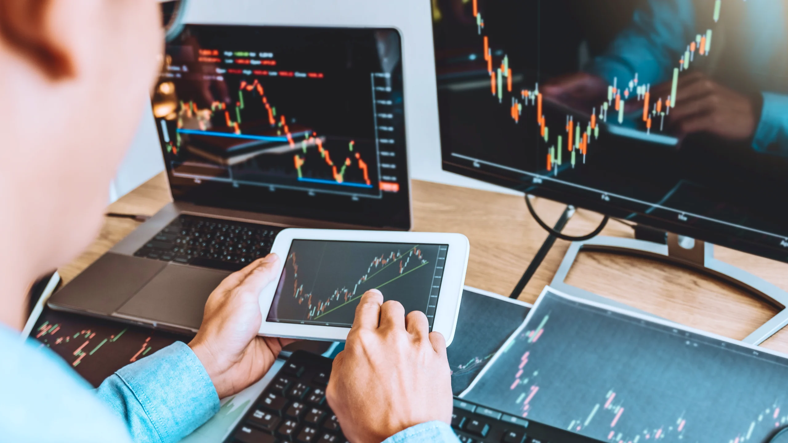 man looking at pad, and his desk is full of papers, computer monitor and laptop, all of these are about the same cfd trading charts