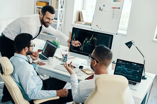 three businessmen engaging in a discussion regarding trading indices shown on the monitor screen, and two laptops