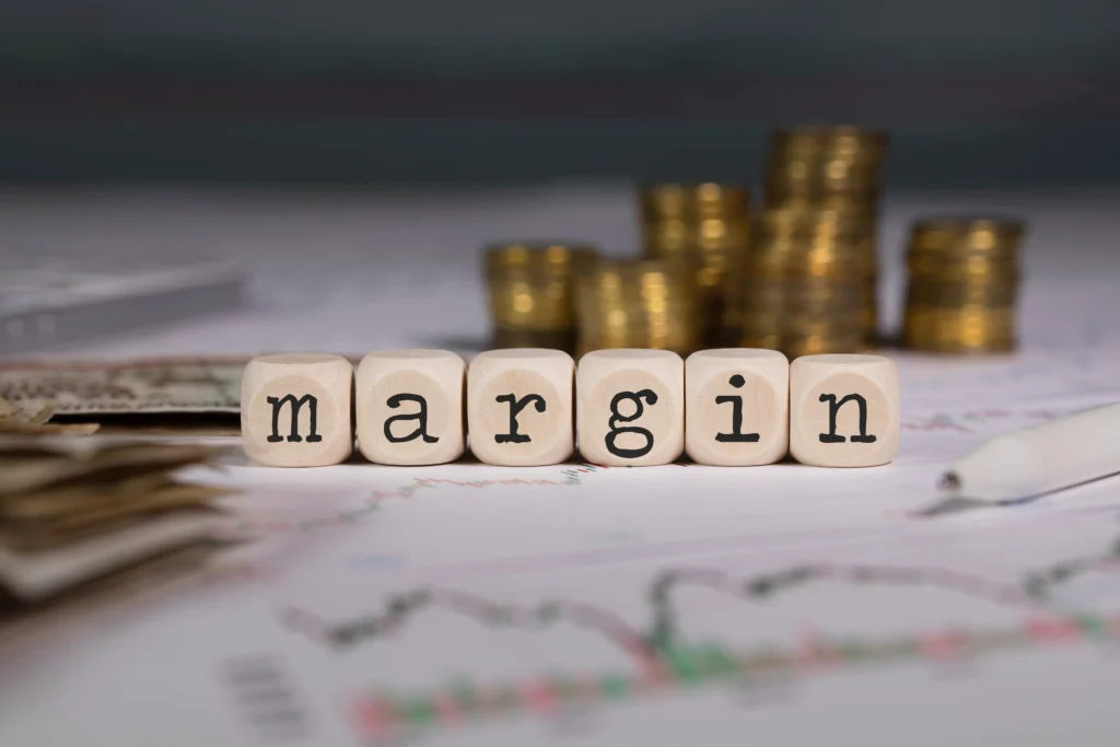 six small wooden cubes printed with ‘margin’ are placed in the background of currency, stock trends, etc.