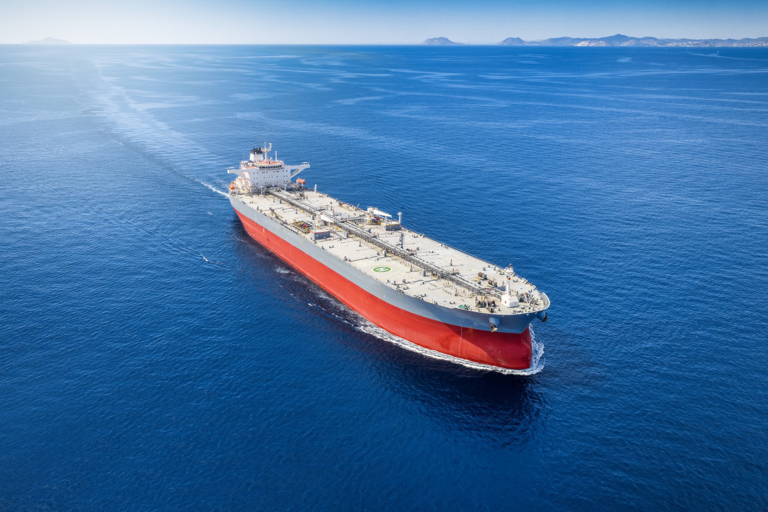 A large red-and-white tanker ships through calm blue ocean with distant islands on the horizon at left and right.