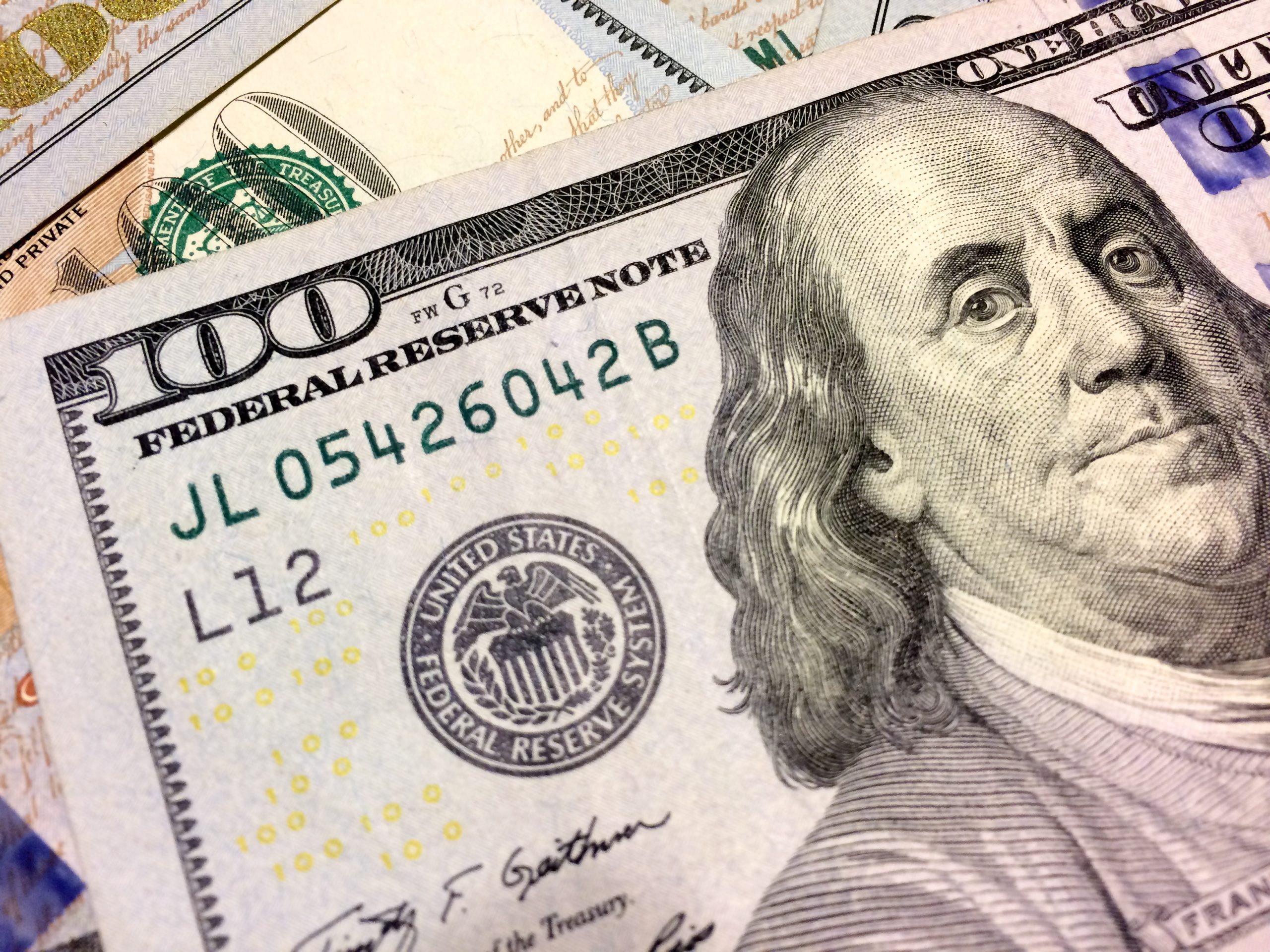 Close-up of overlapping U.S. 100-dollar bills, highlighting the Benjamin Franklin portrait and security features like the green serial numbers and seals.