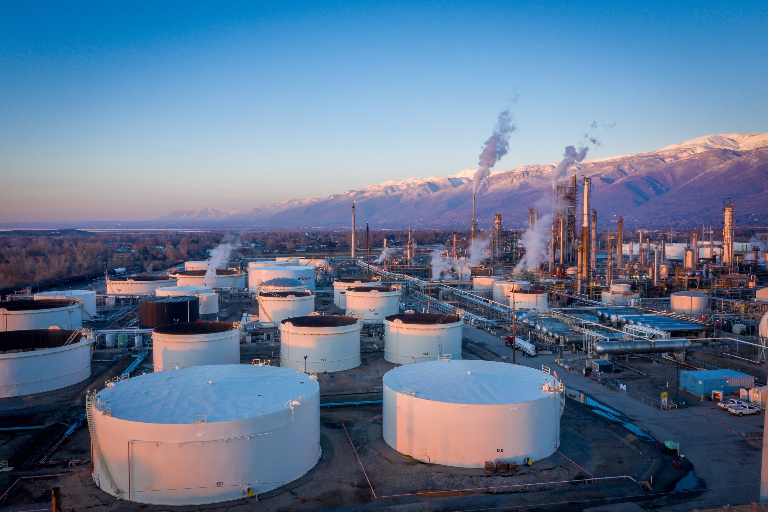 Industrial refinery with many white storage tanks and smokestacks, set against snow-capped mountains at dusk.