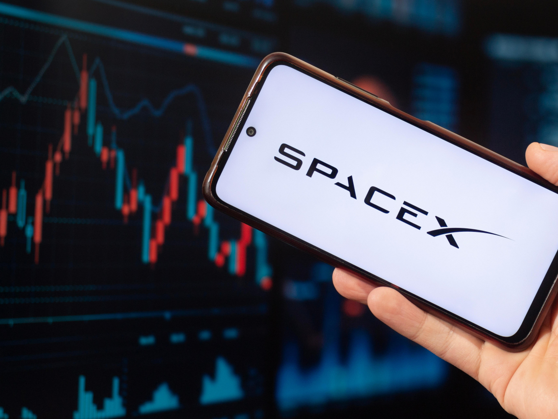 Phone displaying SpaceX logo held in front of a dark financial chart background, implying tech/space investment context.