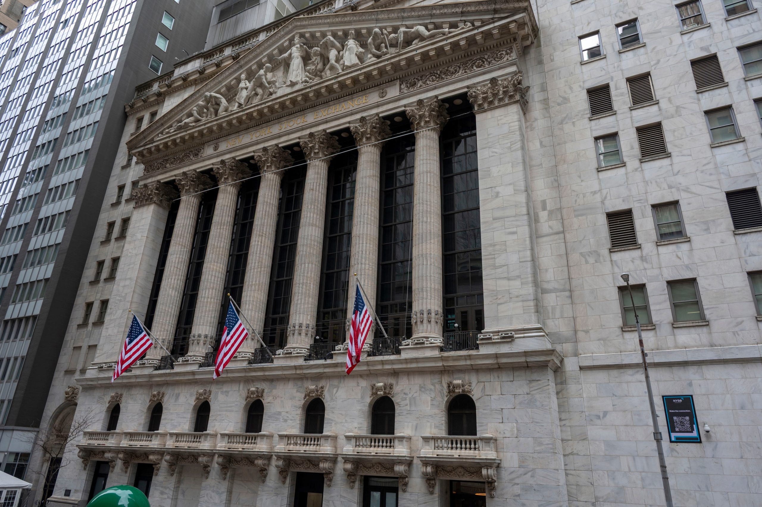 Front view of the New York Stock Exchange building with tall Corinthian columns and gold inscription, flanked by American flags in the foreground.