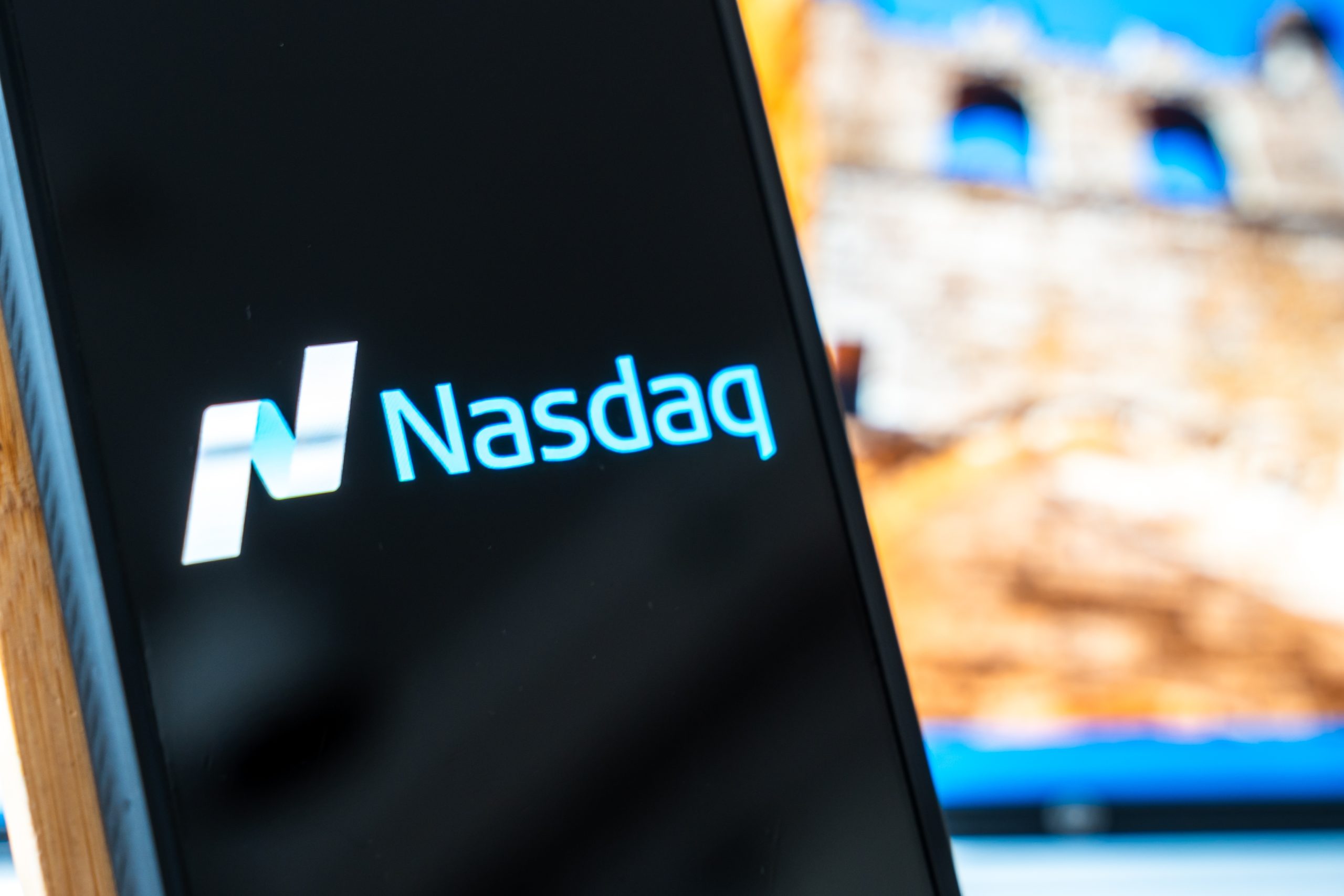 Close-up of a curved device screen displaying the blue NASDAQ logo against a dark background with a blurred outdoor backdrop.
