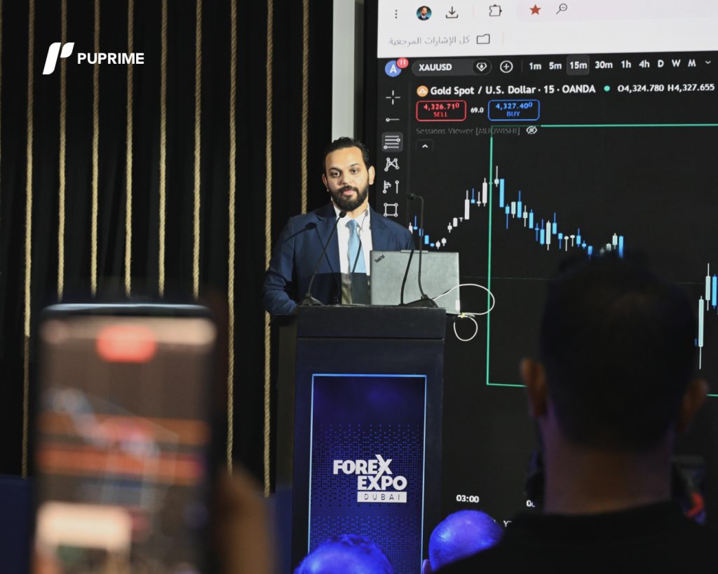 1 1 – PU Prime | More Than Trading Man in a suit presenting at Forex Expo Dubai, standing at a podium with a laptop, large screen showing a stock chart behind him.