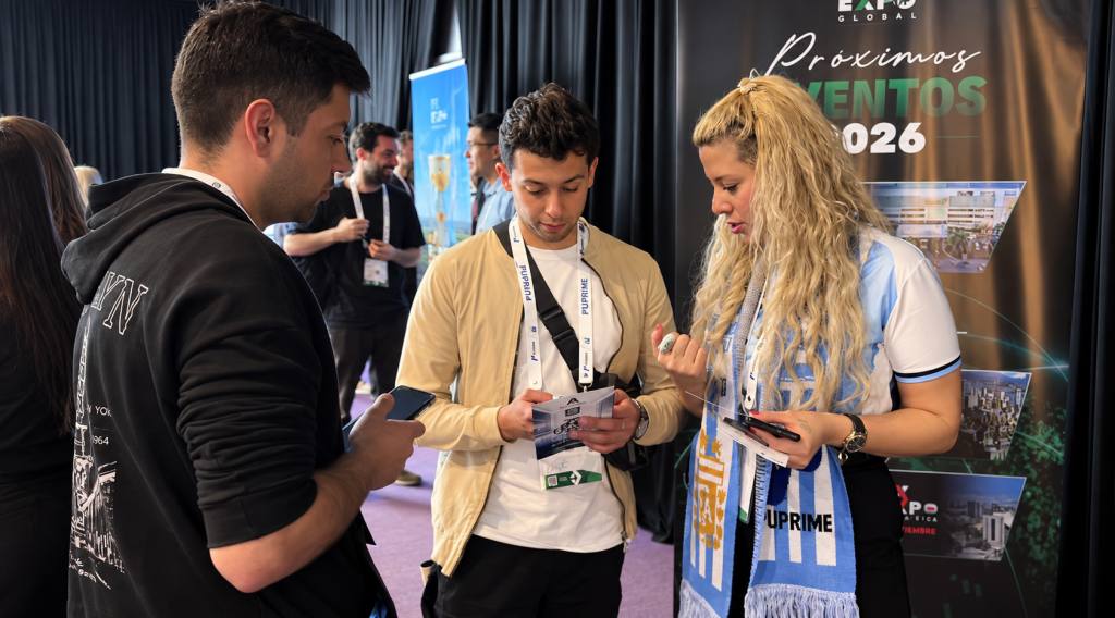 image – PU Prime | More Than Trading Three event attendees review pamphlets at a conference booth with a promotional banner in the background.