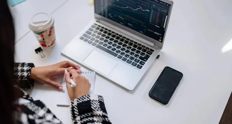 Trader making notes beside a laptop displaying financial charts.