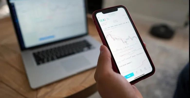 Hand holding a smartphone with a trading chart beside a laptop.