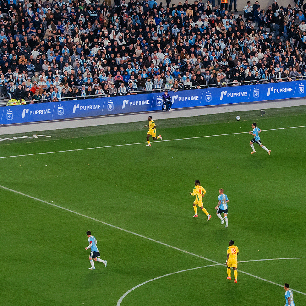 infographic 3 – PU Prime | More Than Trading Soccer match with players in yellow and light-blue jerseys on a green field and a large crowd in the stands behind the sideline banner.