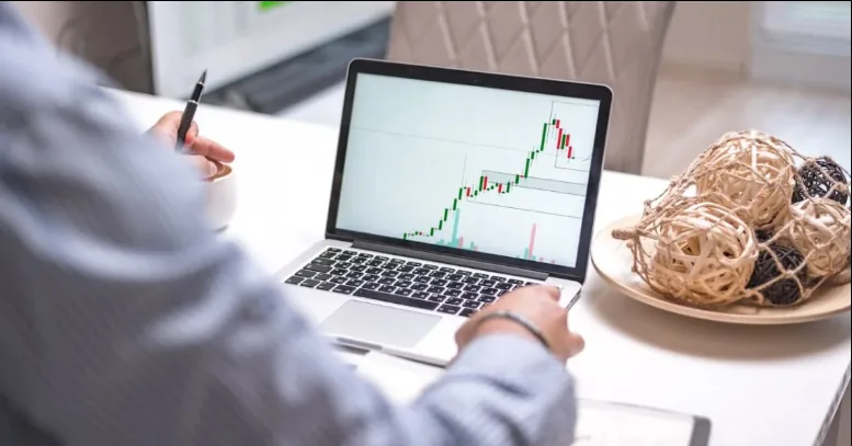 Trader reviewing an upward-trending chart on a laptop with a pen in hand.