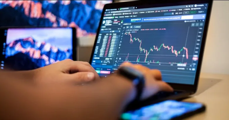 Trader using a laptop with market charts and a smartphone nearby.