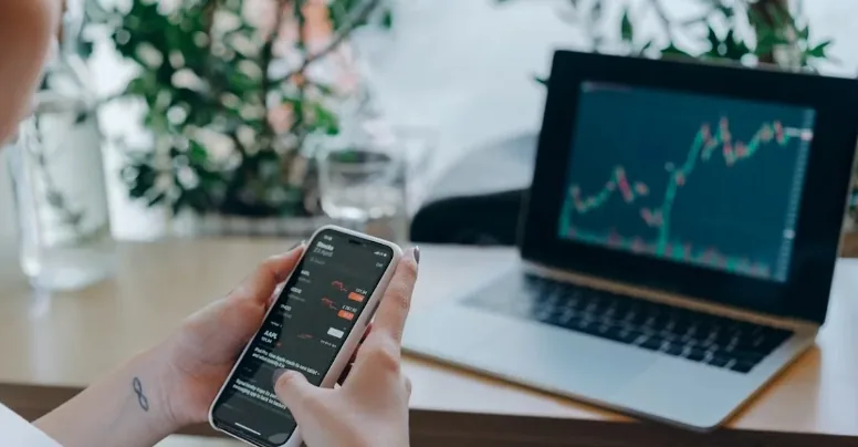 Hand holding a phone with market quotes beside a laptop displaying a price chart.